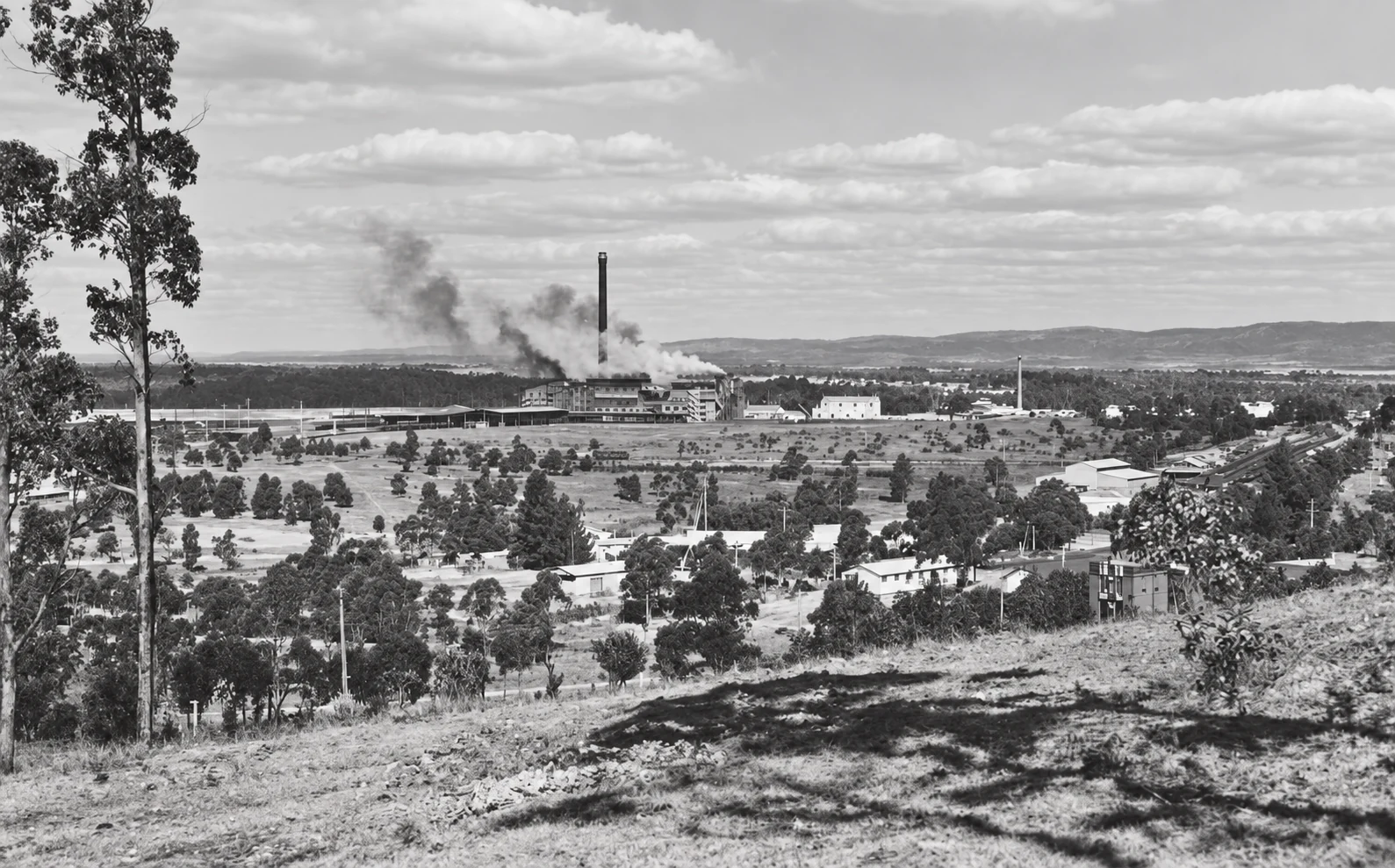 Wide black-and-white view across the Yallourn township landscape towards the briquetting works, chimney and smoke in the middle distance.