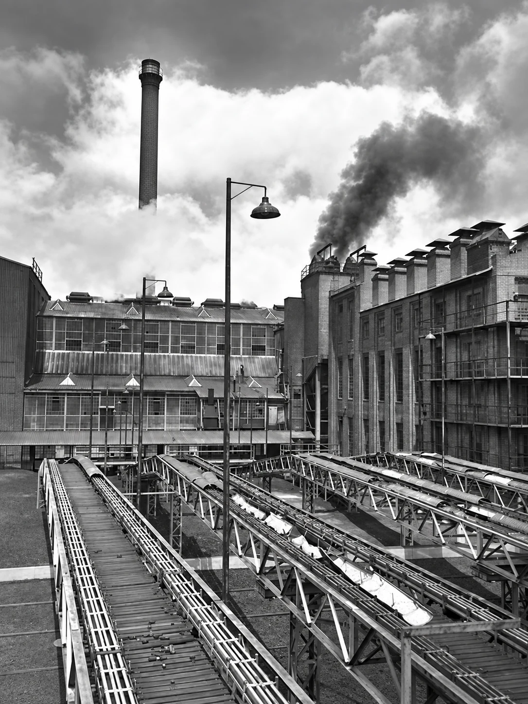 Vertical black-and-white view along multiple elevated conveyors between the industrial buildings of the Yallourn briquette works, with smoke rising behind them.