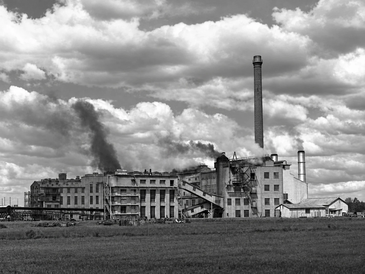 Wide black-and-white view of the Yallourn briquette works beneath a cloud-filled sky, with smoke rising near the factory buildings.