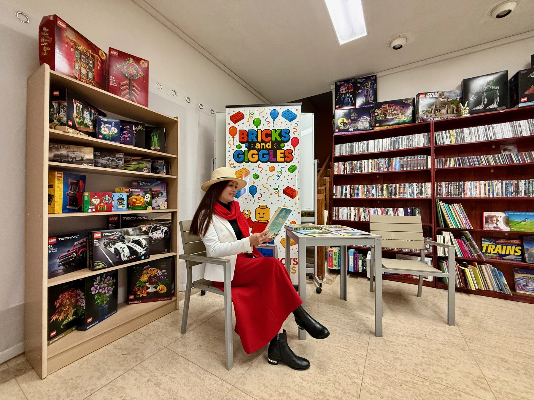 A person in a white jacket, red scarf and red skirt sits reading inside the Yallourn Production Centre among LEGO displays and shelves of books and films.