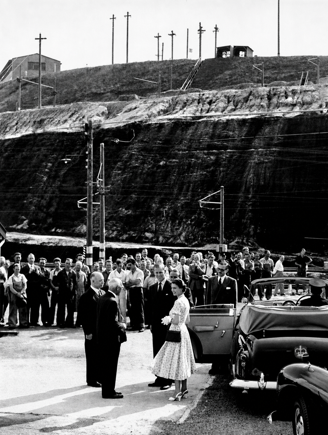 Queen Elizabeth II stands with officials beside two royal cars, with workers and the terraced Yallourn open cut behind them.
