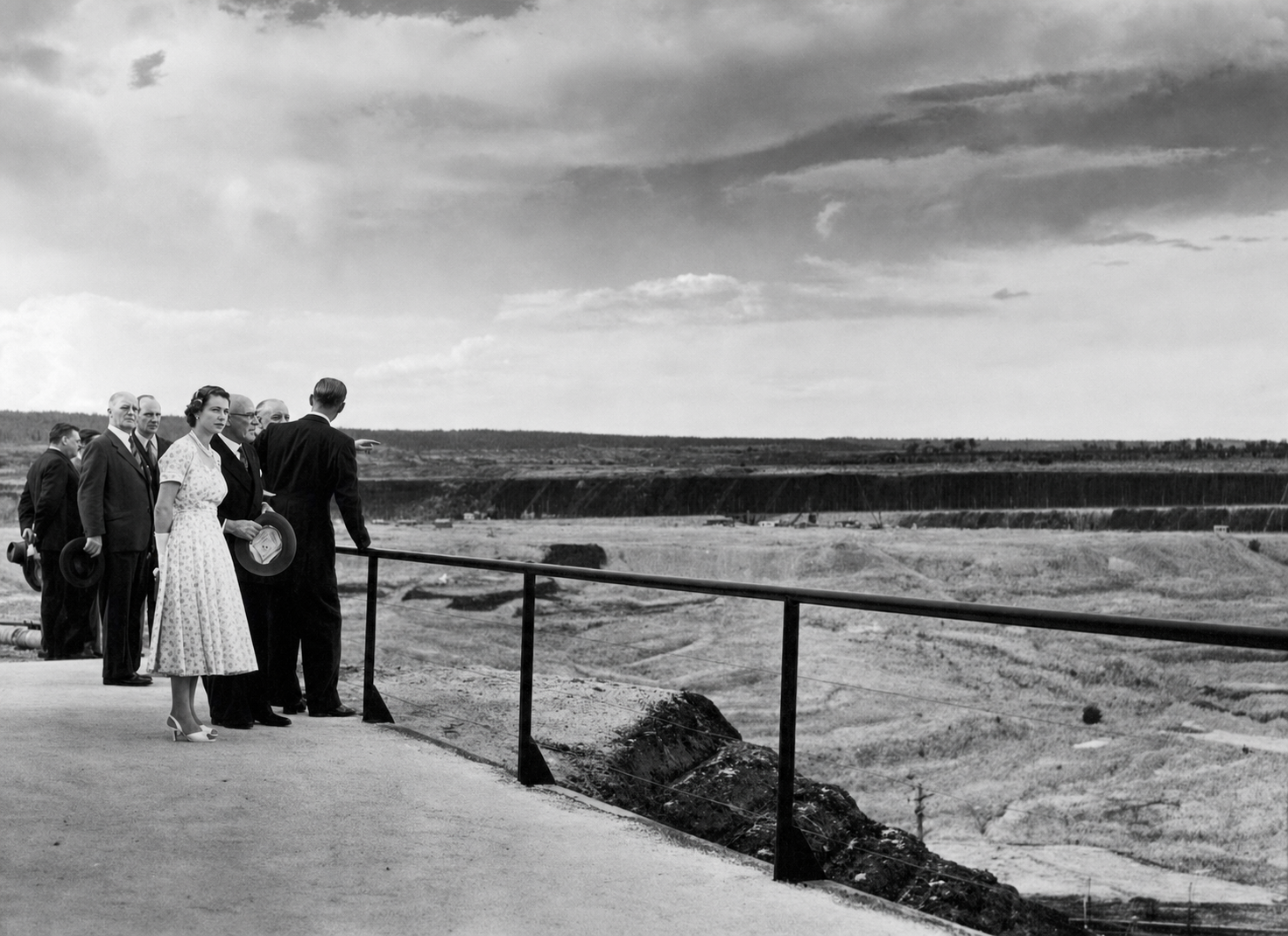 Queen Elizabeth II, the Duke of Edinburgh and accompanying officials stand behind a railing overlooking the broad terraced Yallourn open-cut mine.