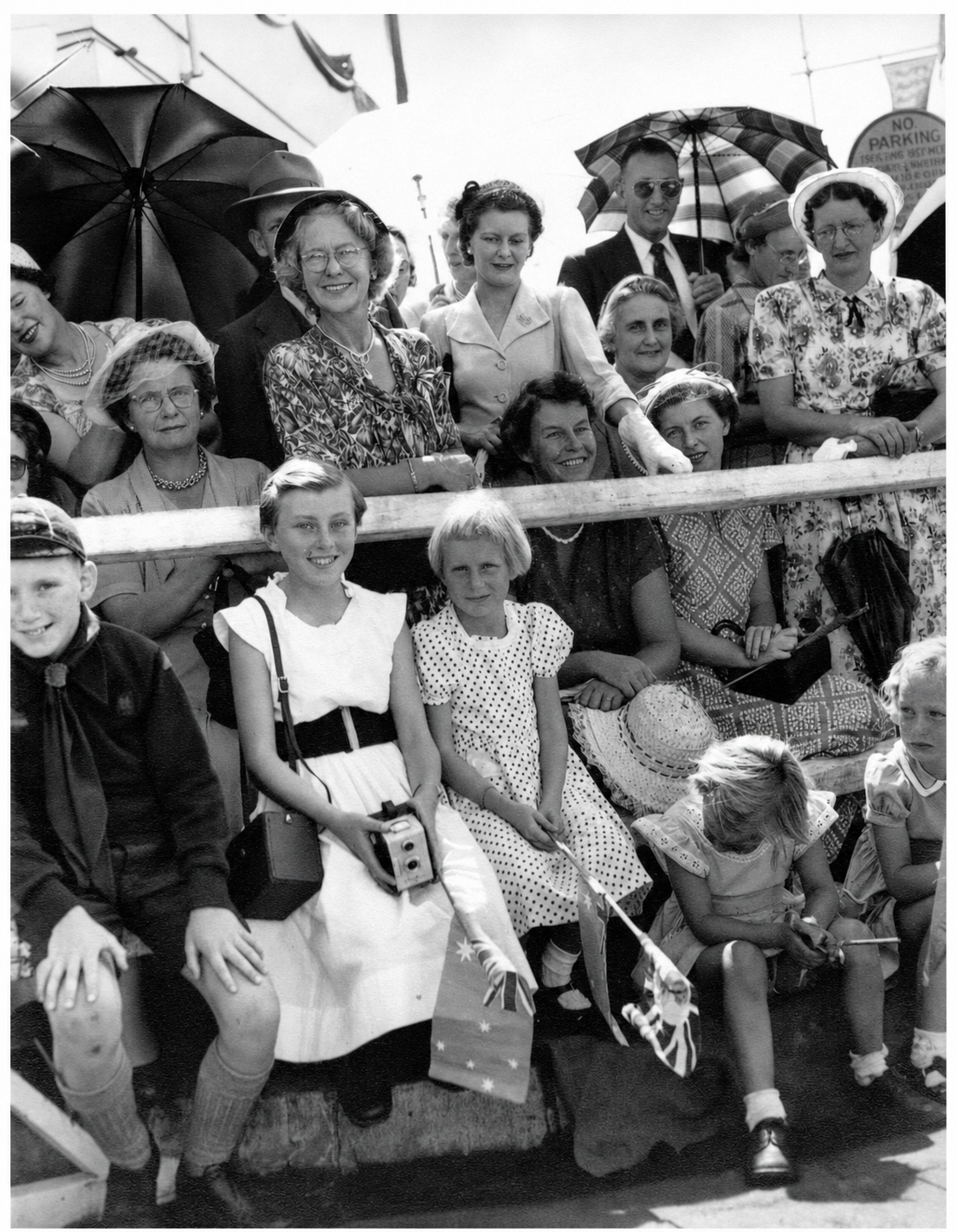 Women and children gather behind a timber rail at Yallourn, some holding umbrellas, cameras and small flags as they wait for the royal procession.