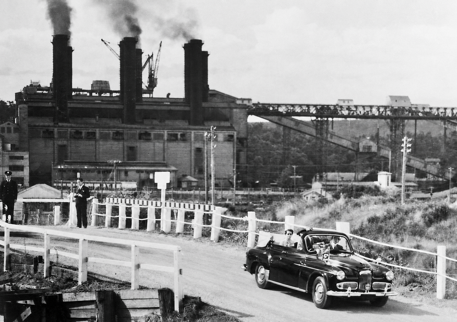 A royal car travels along a fenced road with Yallourn's power station buildings, chimneys and coal conveyors behind it.