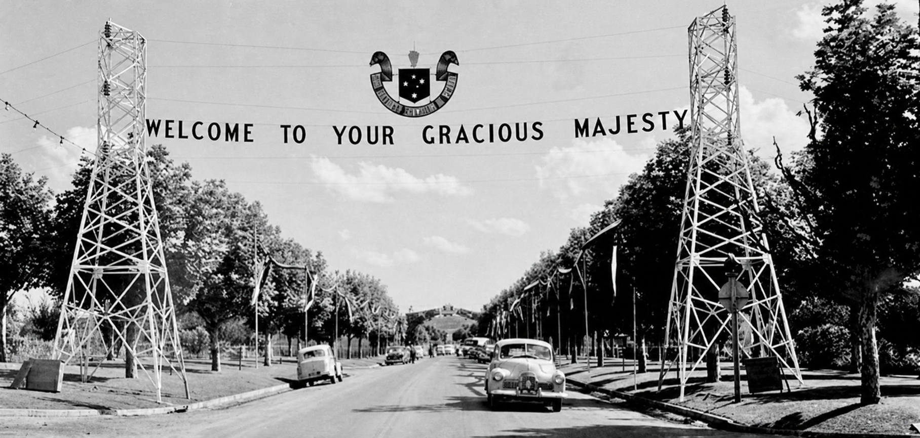 A royal car travels beneath a welcome banner suspended between two lattice towers over Broadway in Yallourn.