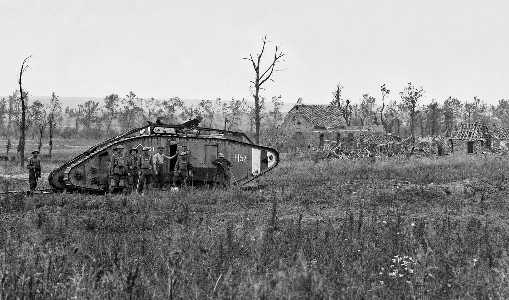 Soldiers stand around a disabled tank marked H52 near buildings and open fields.