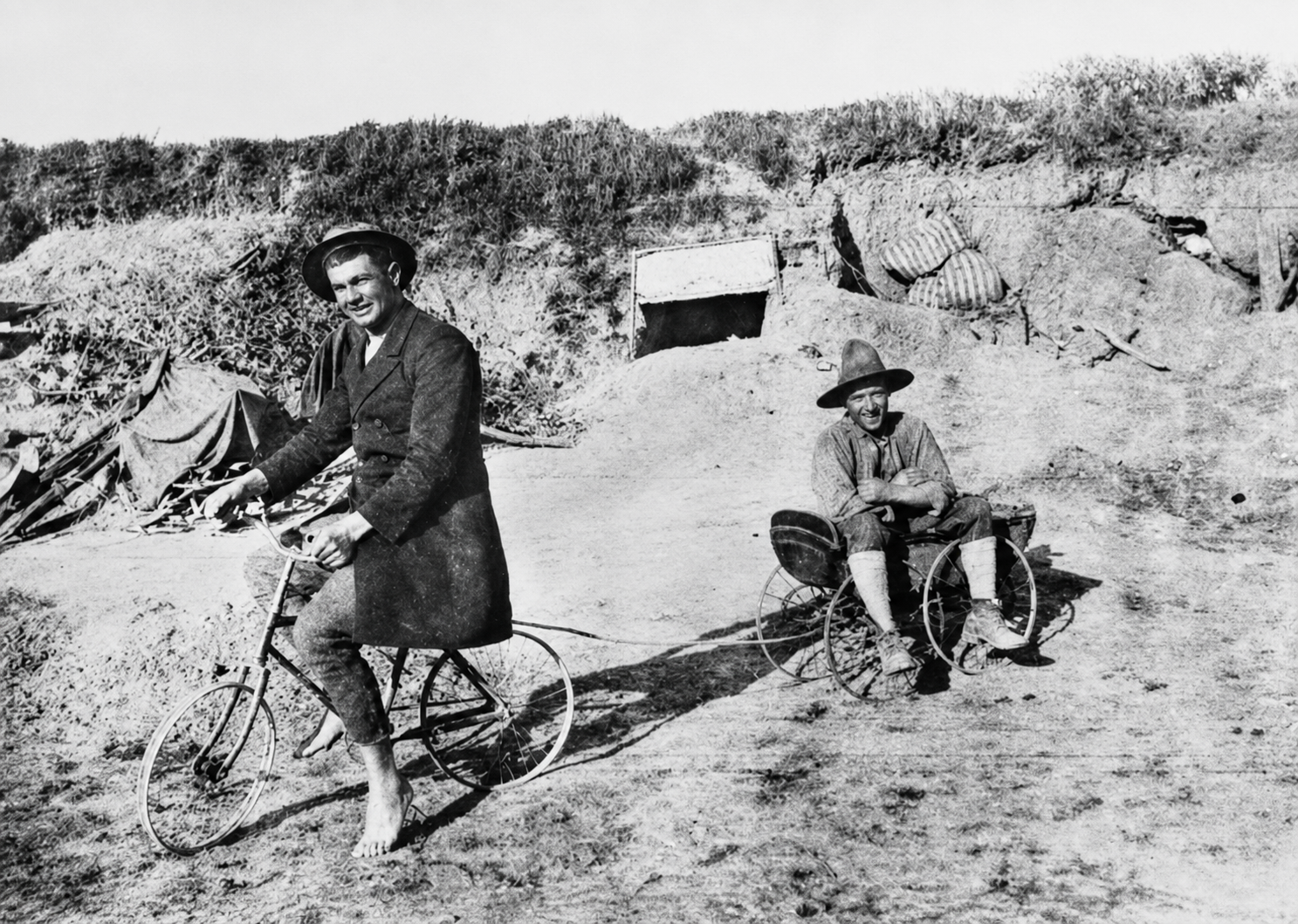 One soldier sits on a small bicycle while another sits in a perambulator beside a dugout.