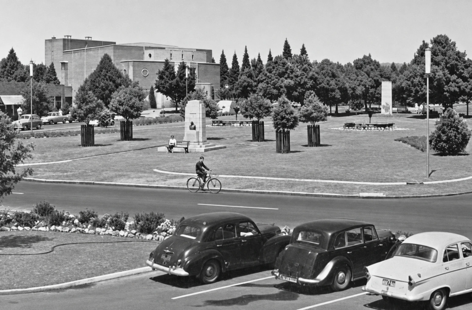 Restored black-and-white wide view across a Yallourn street, with three parked period cars in front and the small Monash memorial, cyclist, lawns, trees and theatre beyond.
