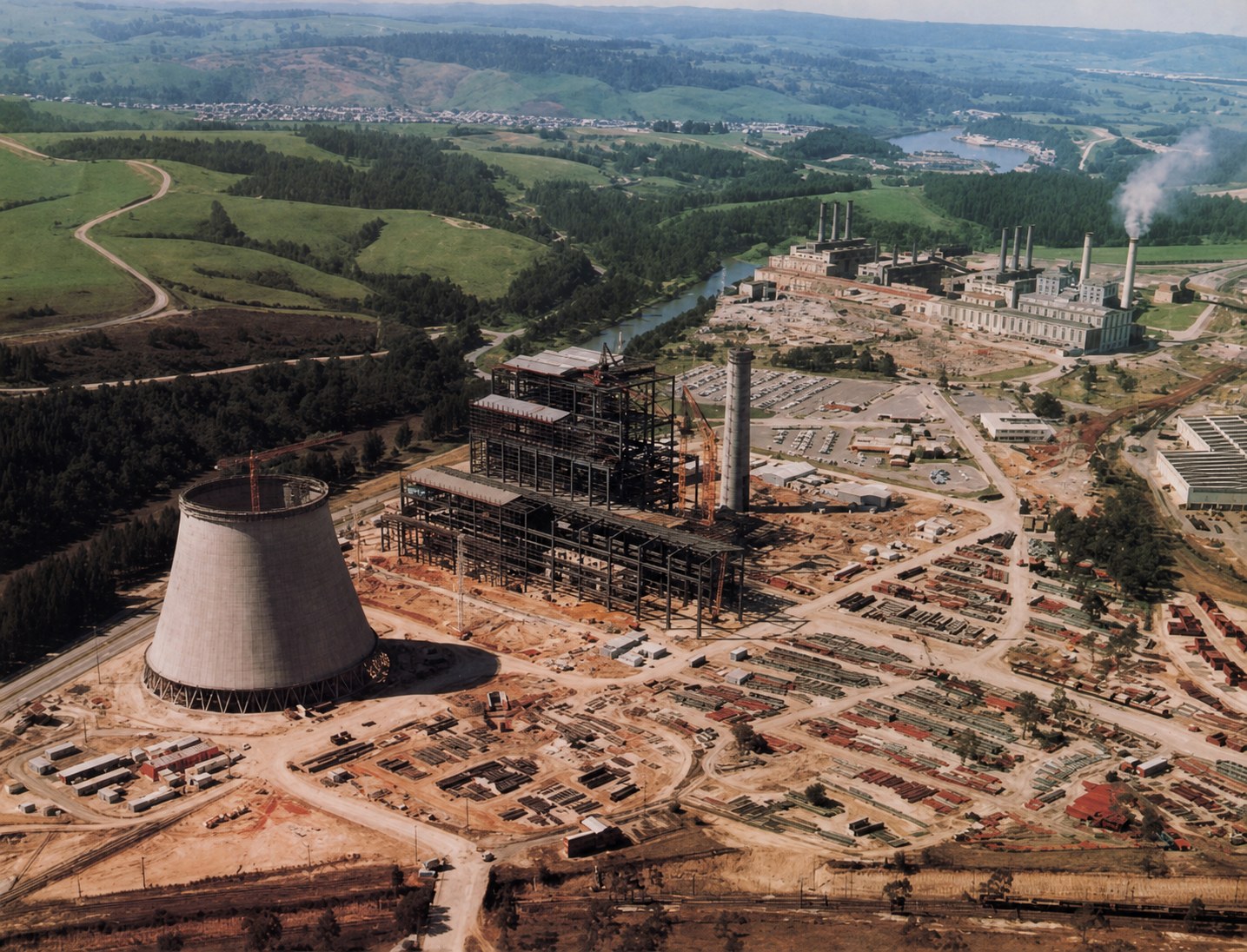 Aerial view of Yallourn W under construction, showing one concrete cooling tower beside the steel-framed station and earlier power-station buildings beyond.