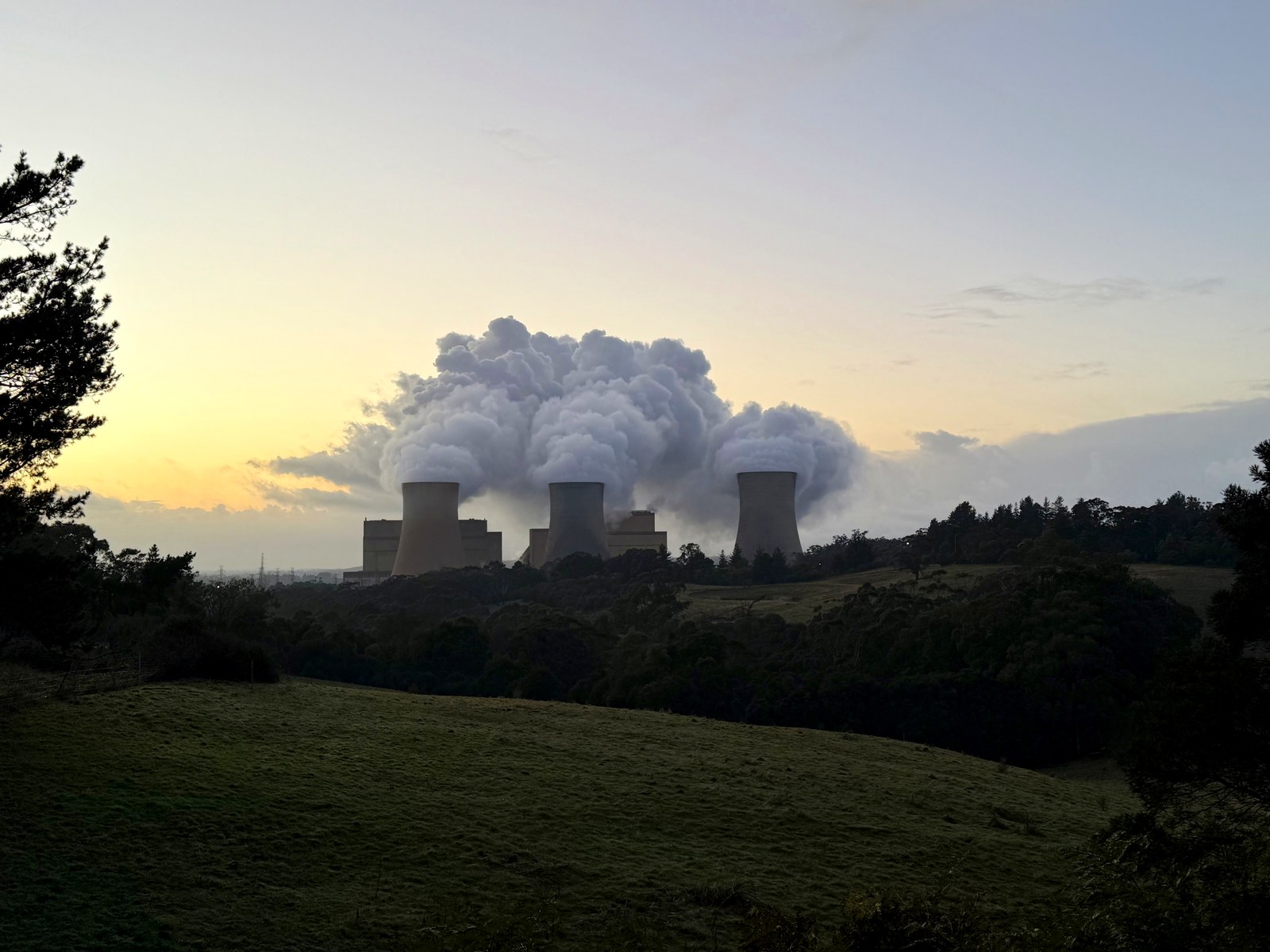 Three Yallourn W cooling towers releasing dense white moisture plumes above a dark wooded landscape at sunrise.