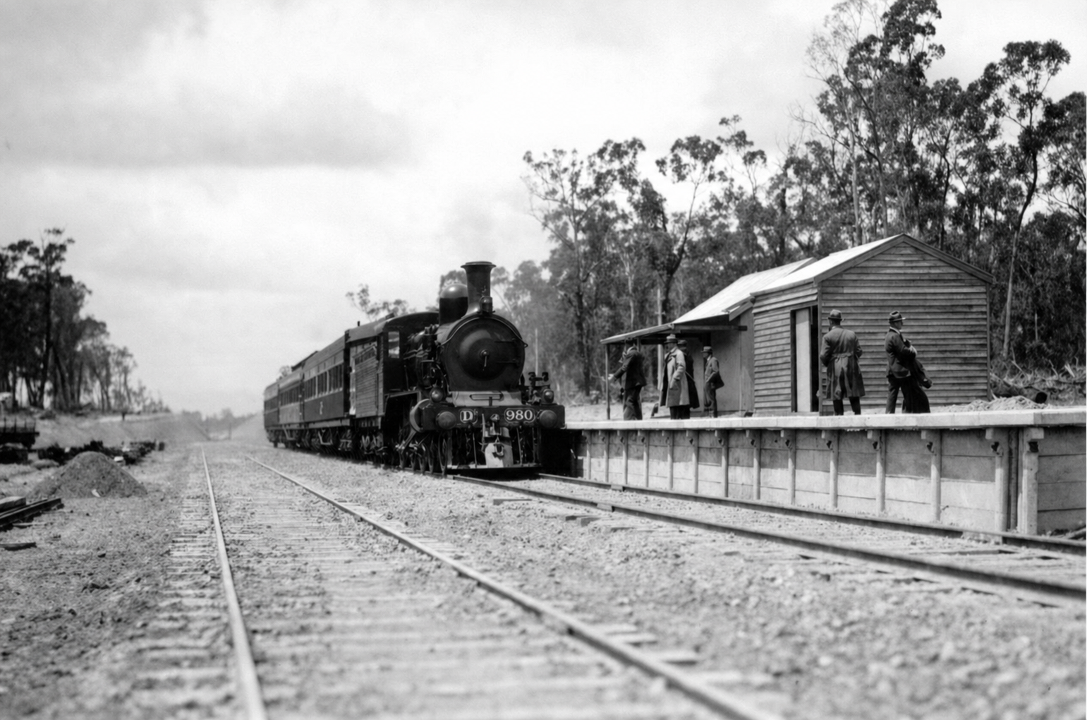 Steam locomotive D 980 and passenger carriages beside a small timber station, with several people standing along the raised platform.