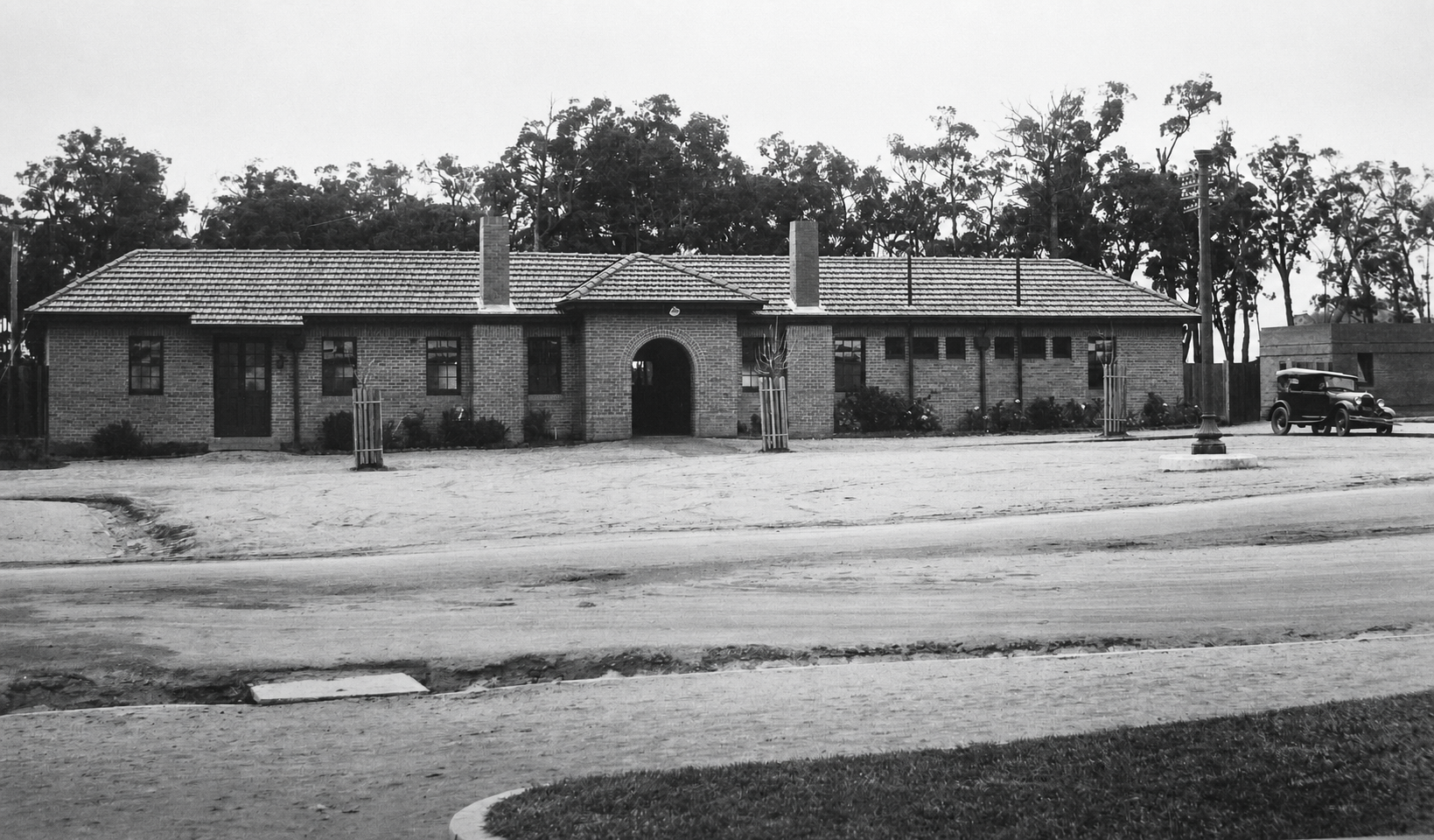 A long single-storey brick railway station with a tiled roof and central arched entrance, viewed across a broad forecourt with young trees and an early motor car.