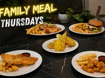 A dark dining table at Morwell RSL set with multiple plates of food including fish and chips, a burger, nuggets and garlic bread under glowing neon signage.