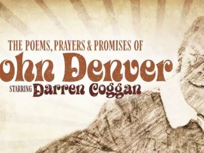 A performer on a warmly lit stage with an acoustic guitar, evoking the folk and country atmosphere of a John Denver tribute concert.