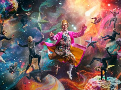 A circus performer in a colourful space-themed costume mid-acrobatic leap, surrounded by projected stars and planets on a darkened stage.