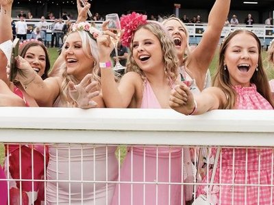 Guests dressed in pink attire gathered at a racecourse luncheon table, with racing action visible in the background.