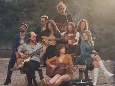 A ensemble of musicians performing on stage in warm golden lighting, evoking the folk and rock sound of 1960s Laurel Canyon.