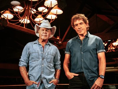 Ian Moss and Troy Cassar-Daley seated on stage together, each holding an acoustic guitar, performing to a theatre audience.