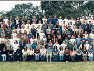 A group of smiling adults at a casual hotel venue, dressed for a school reunion, raising glasses in celebration.