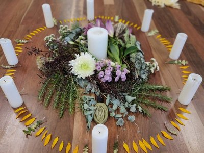 A circle of women sitting together in a warmly lit space, with candles, flowers and a medicine drum at the centre.