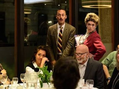 Actors playing Basil Fawlty, Manuel and Sybil in character interacting with laughing guests seated at dinner tables in a decorated event hall.
