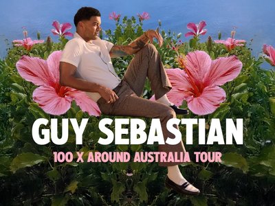 Guy Sebastian performing live on stage with his band, microphone in hand, in front of a large concert audience.