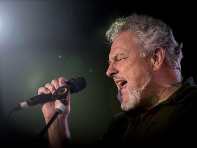 Ross Wilson performing live on stage with his band The Peaceniks, microphone in hand, in front of an energetic crowd.