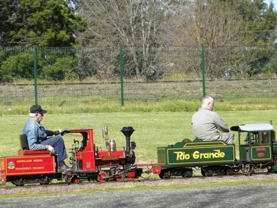 A small steam or diesel miniature train carrying passengers along a track through Newman Park, Traralgon, on a sunny afternoon.