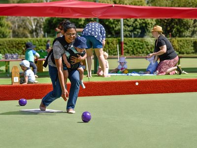 A mother with a pram on a lawn bowls green, laughing with other parents on a sunny morning.