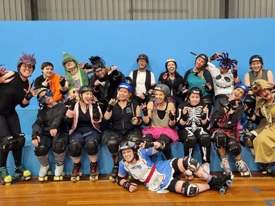 A group of women of mixed skating ability laughing and skating together on a roller derby rink, wearing helmets and protective gear.