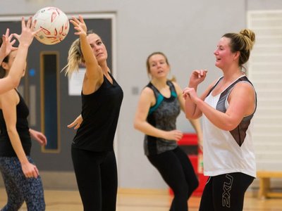 A group of women of mixed ages playing walking netball on an indoor court, smiling and passing the ball.