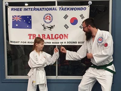 Girls and young women in white taekwondo uniforms practising kicks and stances together in a community hall.