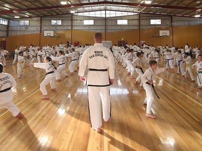 Girls in white taekwondo uniforms practising kicks in a community hall setting.