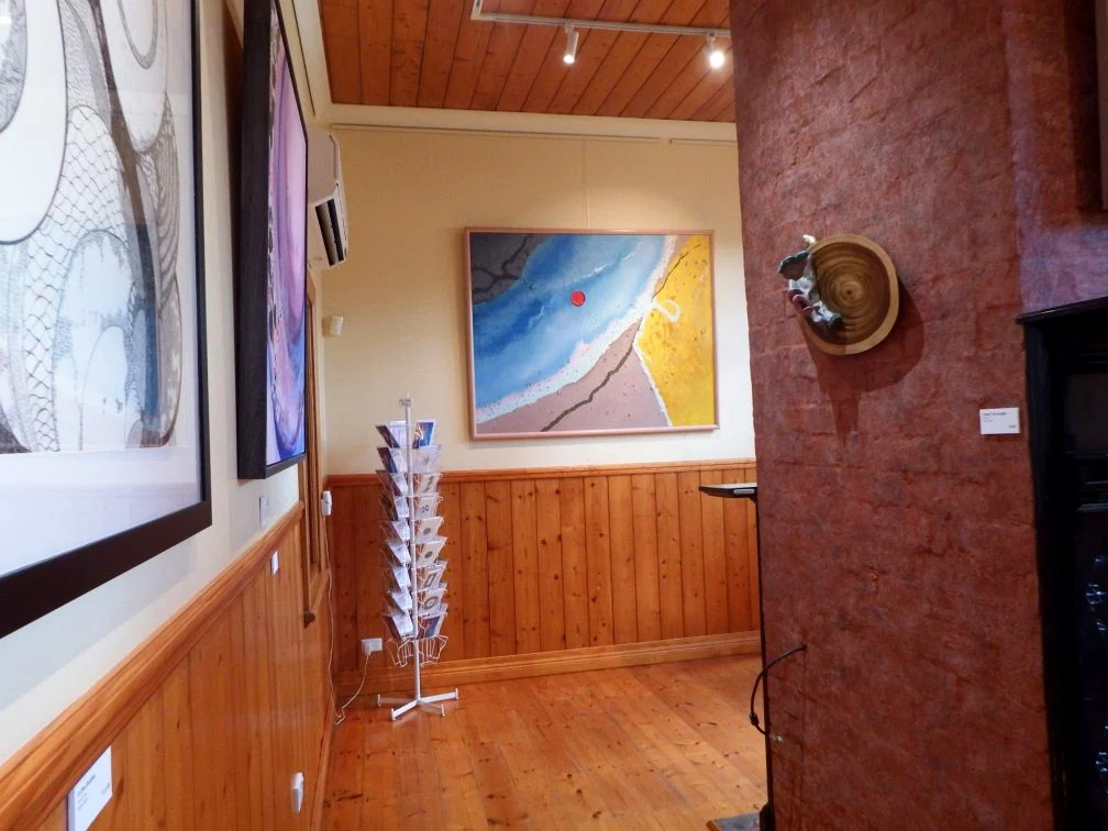 A view through Matchbox Gallery past framed artworks, timber panelling, a display rack and a textured red wall.