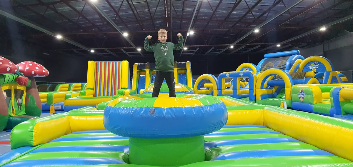 A child standing on a circular inflatable challenge with obstacle courses behind him inside Stadium 34.
