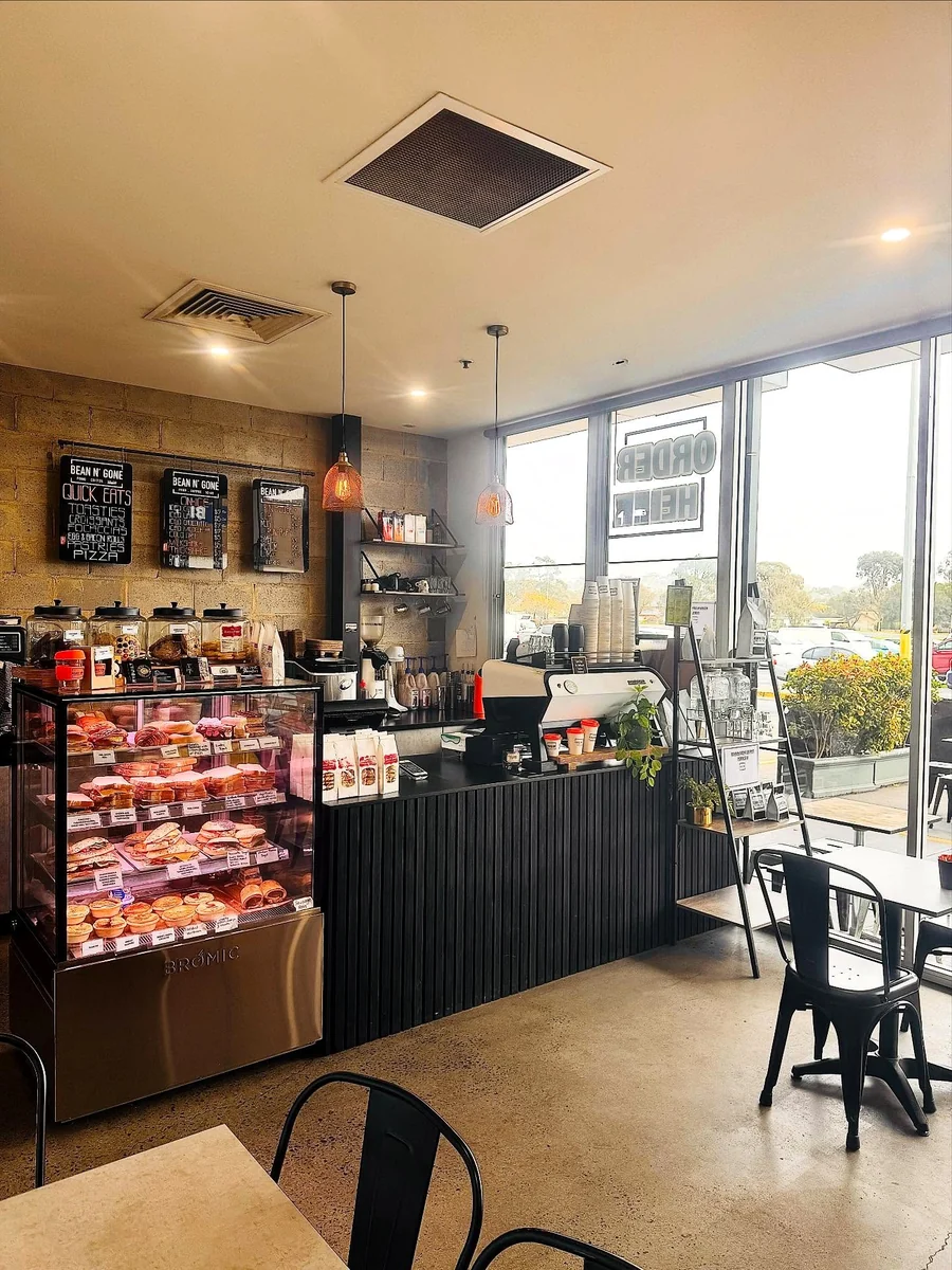 The Bean n' Gone interior with a food cabinet and coffee counter on the left and tables beside full-height windows on the right.