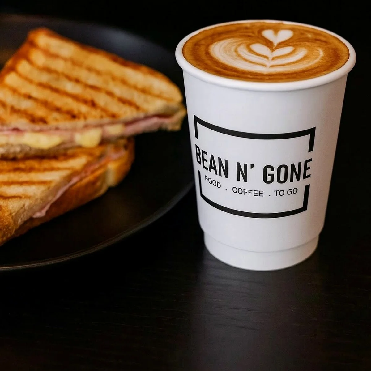 A takeaway coffee with leaf-shaped latte art beside a toasted sandwich on a dark table.