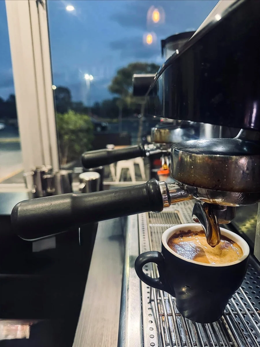Espresso pours into a black cup from a coffee machine beside a window showing blue early-morning light.