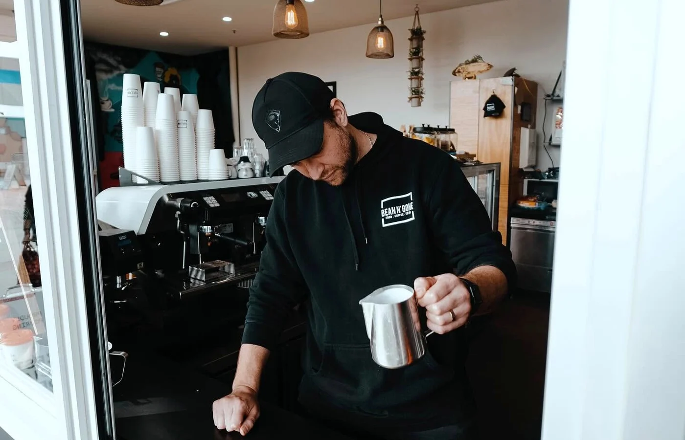 Dean works at the Bean n' Gone coffee window with a cup on the counter and a milk jug in hand.