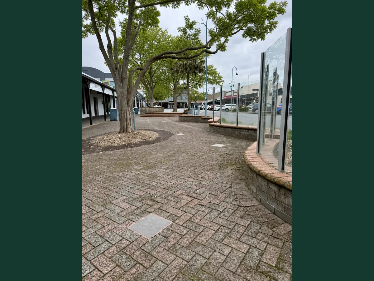 AI-assisted summer seasonal visualisation showing the brick-paved Moore Street footpath, embedded plaques and mature trees in leaf.