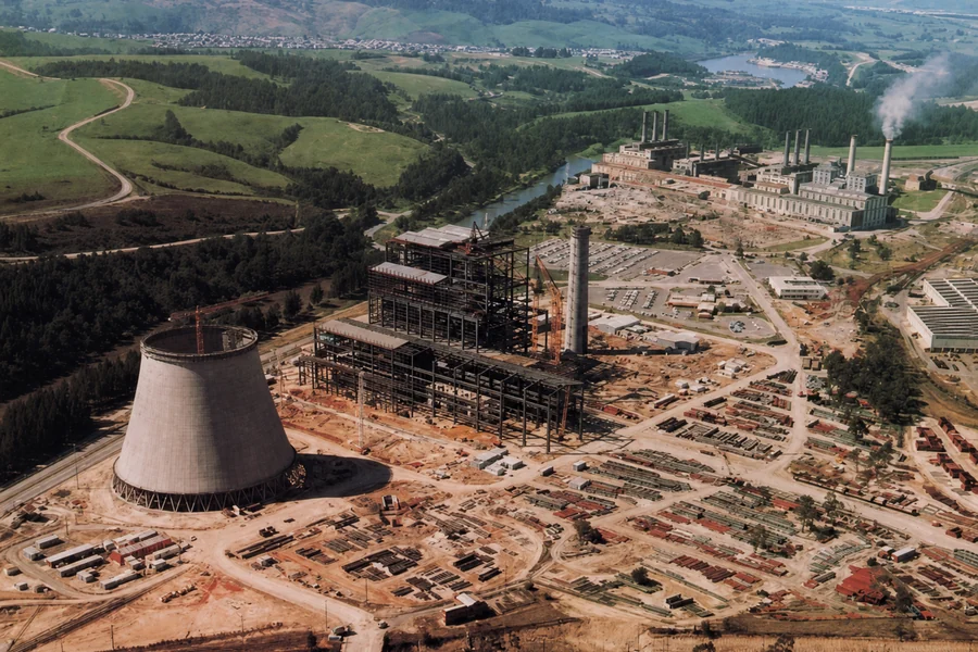 Aerial view of Yallourn W under construction, showing one concrete cooling tower beside the steel-framed station and earlier power-station buildings beyond.