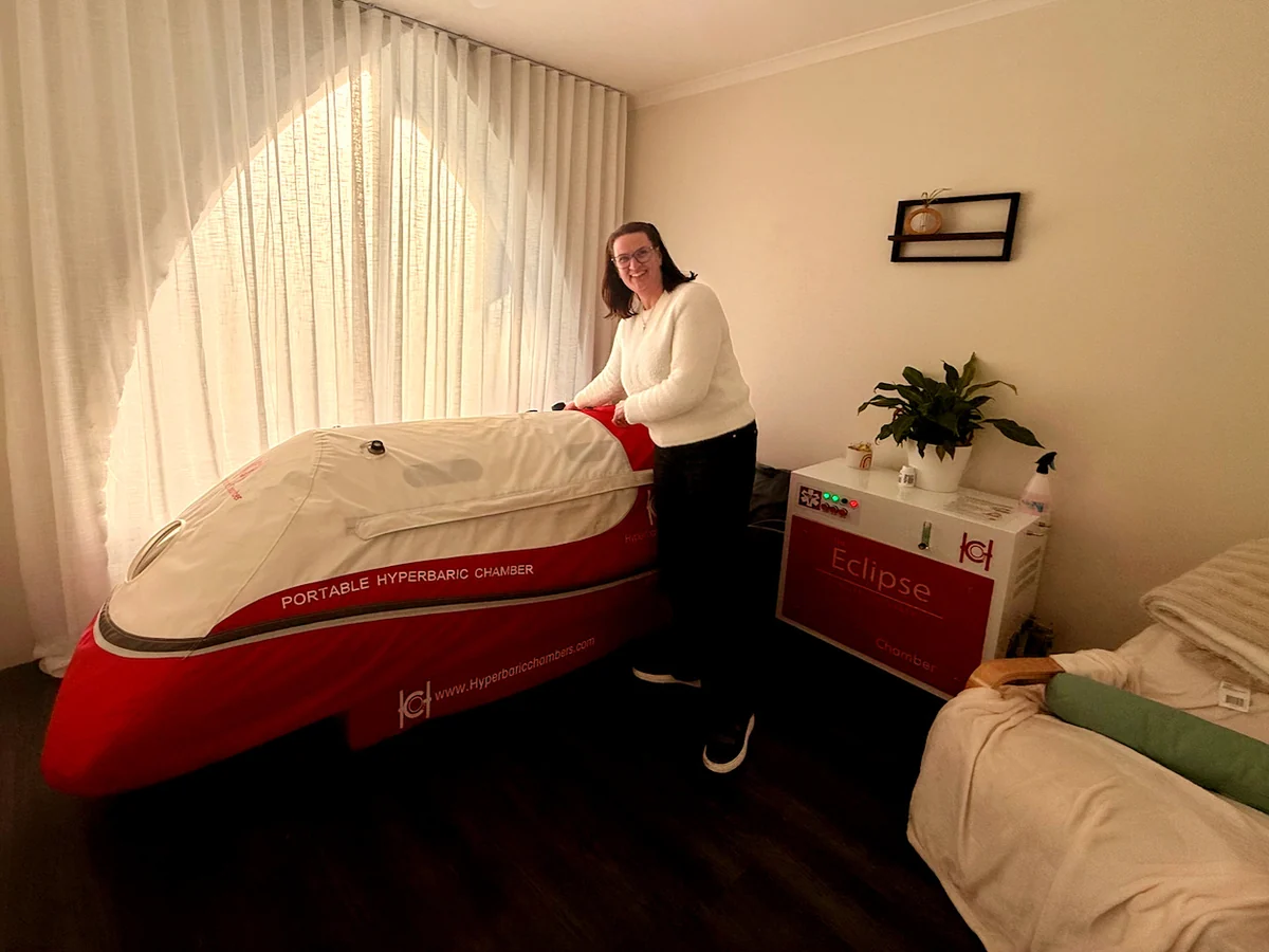 Wendy Stanway stands beside a red and cream portable chamber in a softly lit treatment room.
