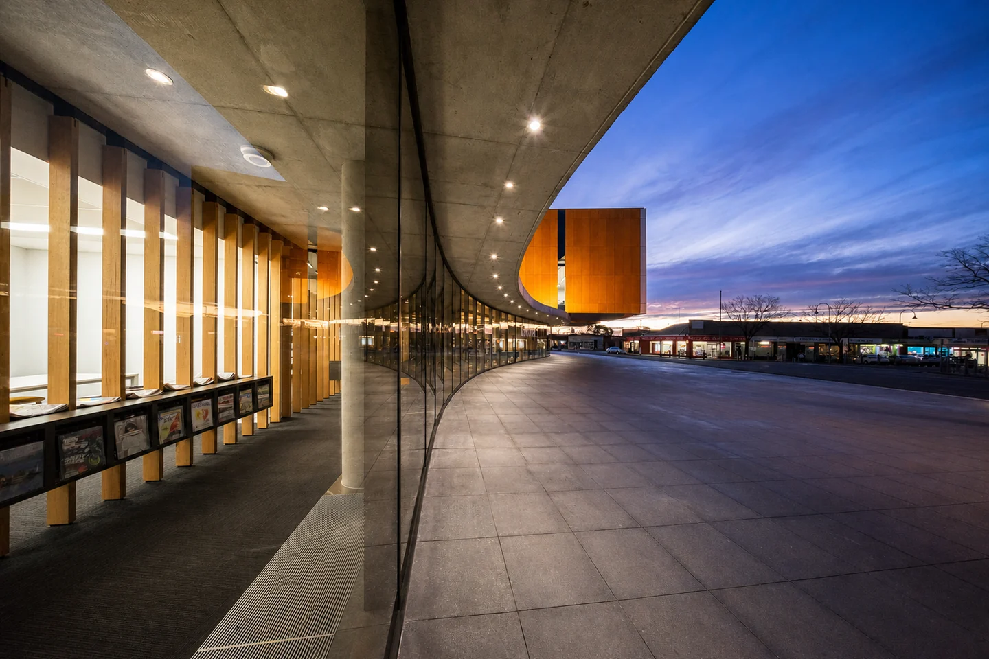 Moe Library's long curved glass facade and timber-lit interior beside the civic plaza at dusk.