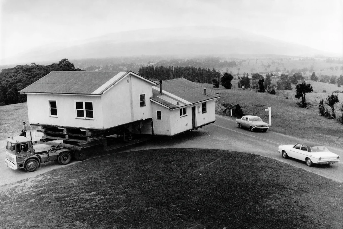 Black-and-white photograph of a Yallourn house carried on a truck along a country road while two period cars wait nearby.