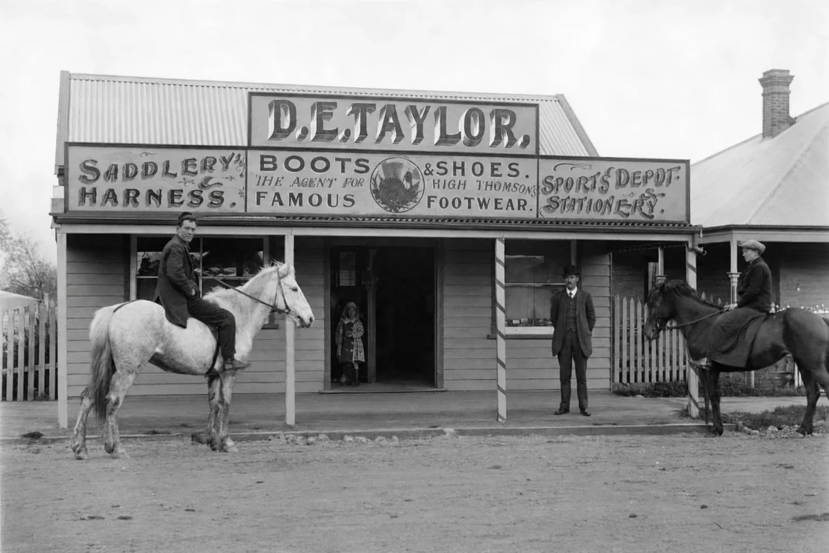 Two mounted men and another man stand outside D. E. Taylor's saddlery, boots and shoes shop in Yinnar around 1930.