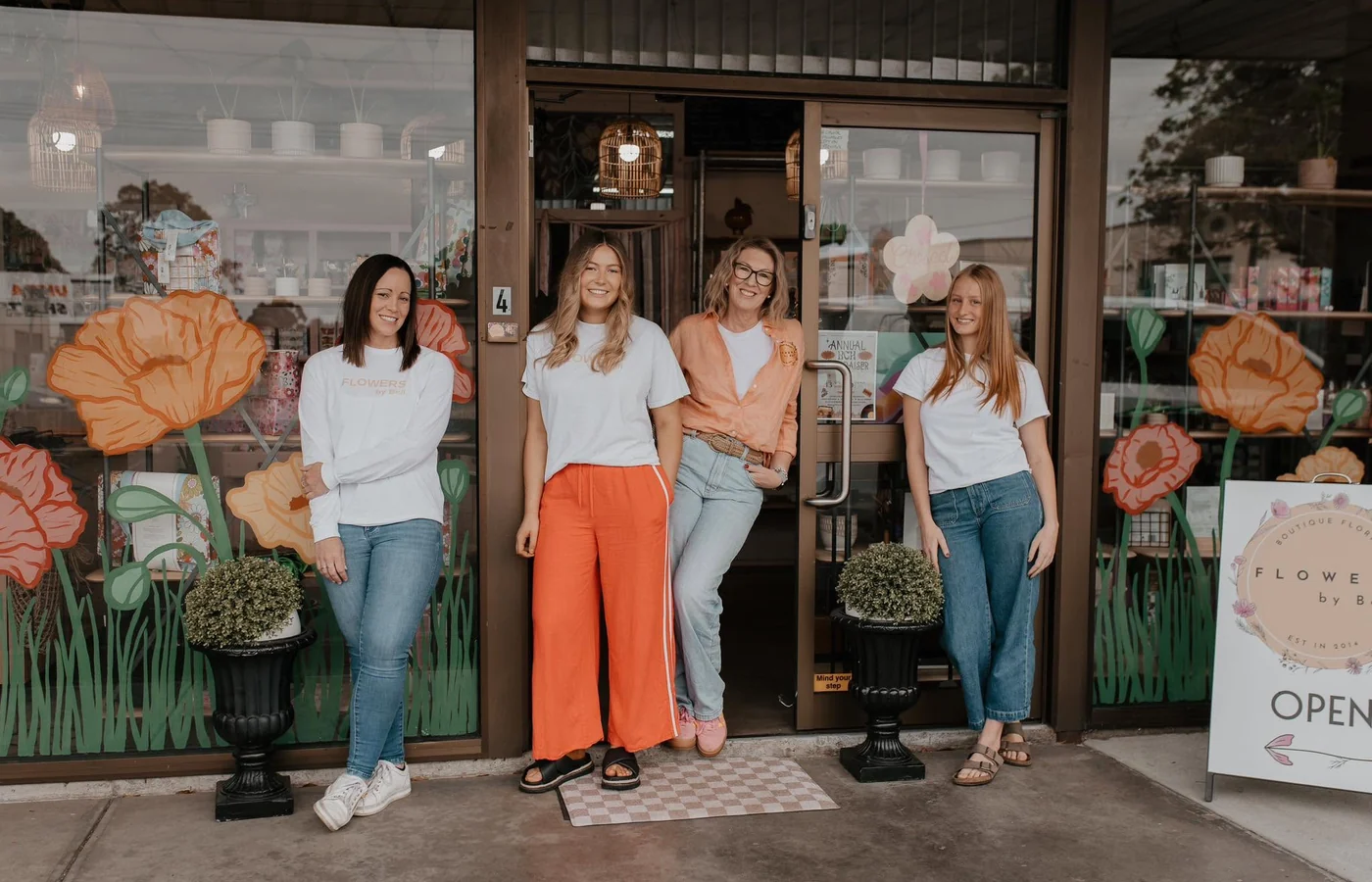 Four Flowers by Bell team members stand outside the Traralgon shopfront, framed by large orange flowers painted across the windows.