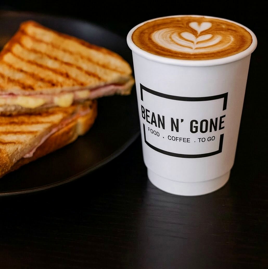 A takeaway coffee with leaf-shaped latte art beside a toasted sandwich on a dark table.