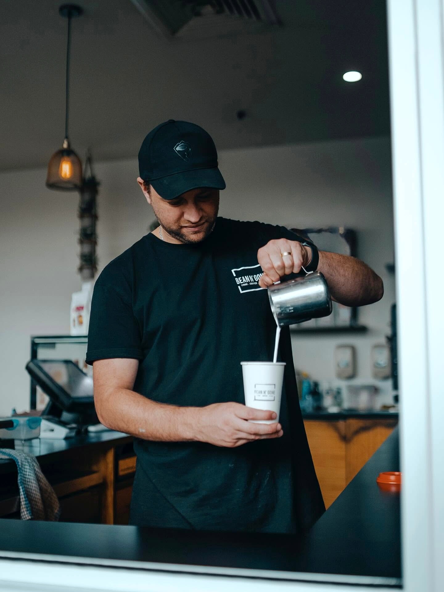 A Bean n' Gone barista pours milk from a metal jug into a takeaway cup at the service window.