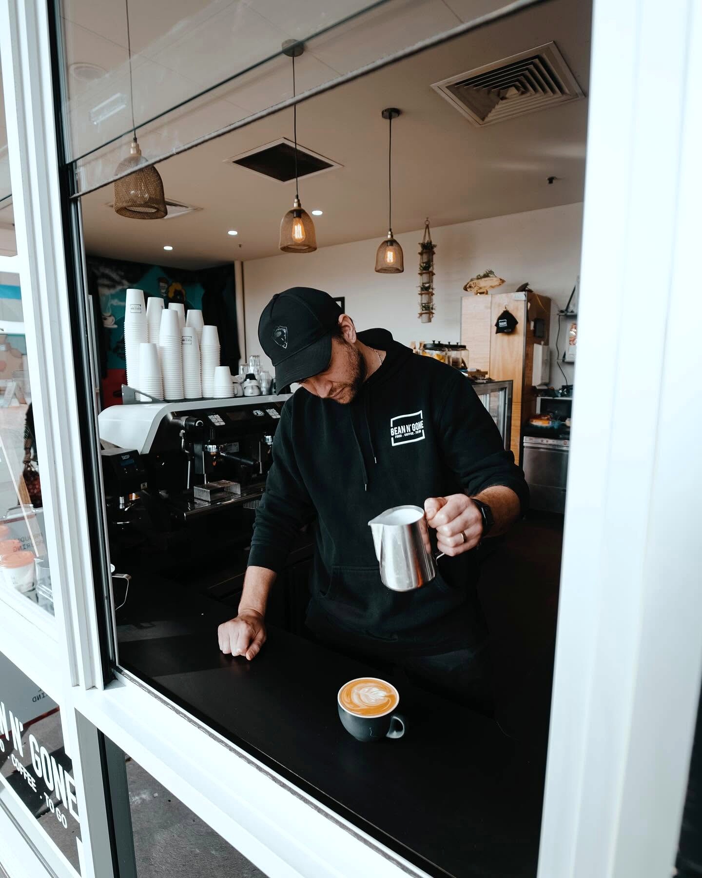 A Bean n' Gone barista works at the coffee window with a cup on the counter and a milk jug in hand.
