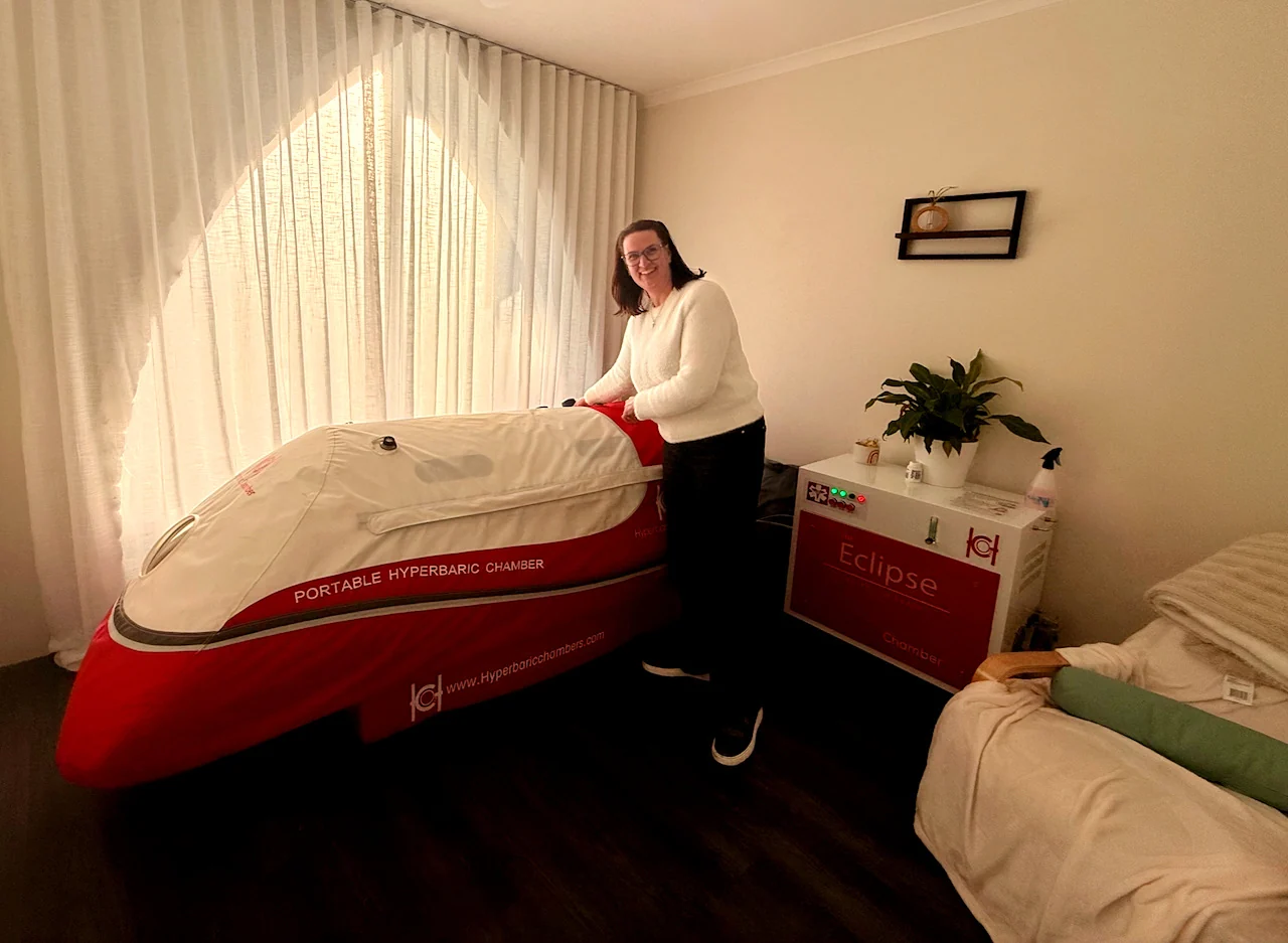 Wendy Stanway stands beside a red and cream portable chamber in a softly lit treatment room.